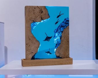 May include: A rectangular wooden art piece featuring a vibrant blue resin scene with manta rays and divers. The wooden frame has a natural, light brown grain. The artwork is set on a wooden base, creating a unique decorative item.
