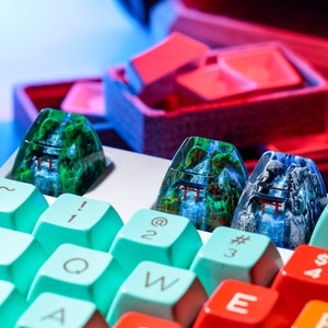 May include: Close-up of custom keyboard keycaps featuring a mountain and waterfall design with a red torii gate. The keycaps are set against a backdrop of colorful keyboard keys in teal, red, and orange. The keycaps are a unique accessory.
