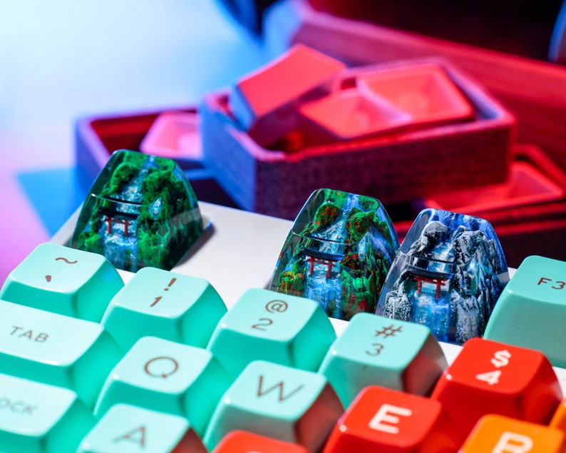 May include: Close-up of custom keyboard keycaps featuring a mountain and waterfall design with a red torii gate. The keycaps are set against a backdrop of colorful keyboard keys in teal, red, and orange. The keycaps are a unique accessory.