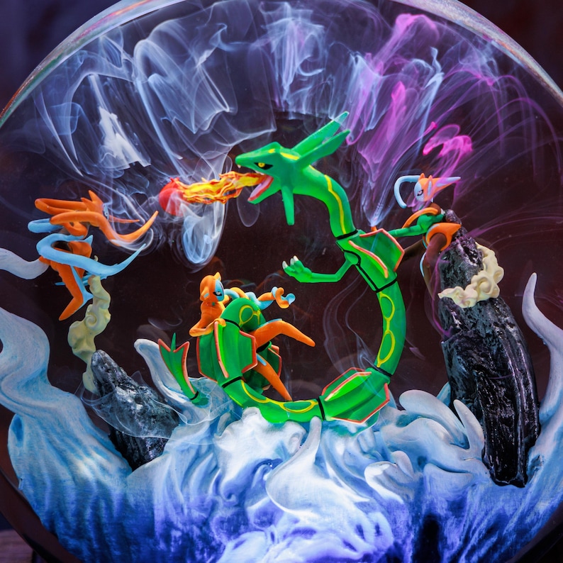 May include: A circular display featuring a green serpentine creature breathing fire, accompanied by orange and blue figures. The scene is set against a backdrop of swirling white and purple smoke effects, with dark rock formations and blue base elements.