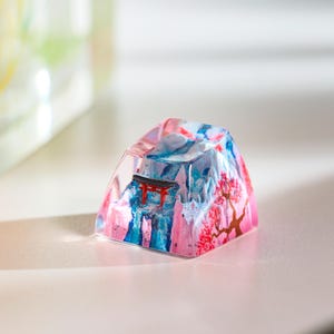 Puede incluir: Una tecla decorativa con un dise&ntilde;o de inspiraci&oacute;n japonesa. Presenta una escena de monta&ntilde;a con una puerta torii roja y flores de cerezo. La tecla est&aacute; hecha de resina y es una mezcla de rosa, azul y blanco. El dise&ntilde;o es art&iacute;stico.