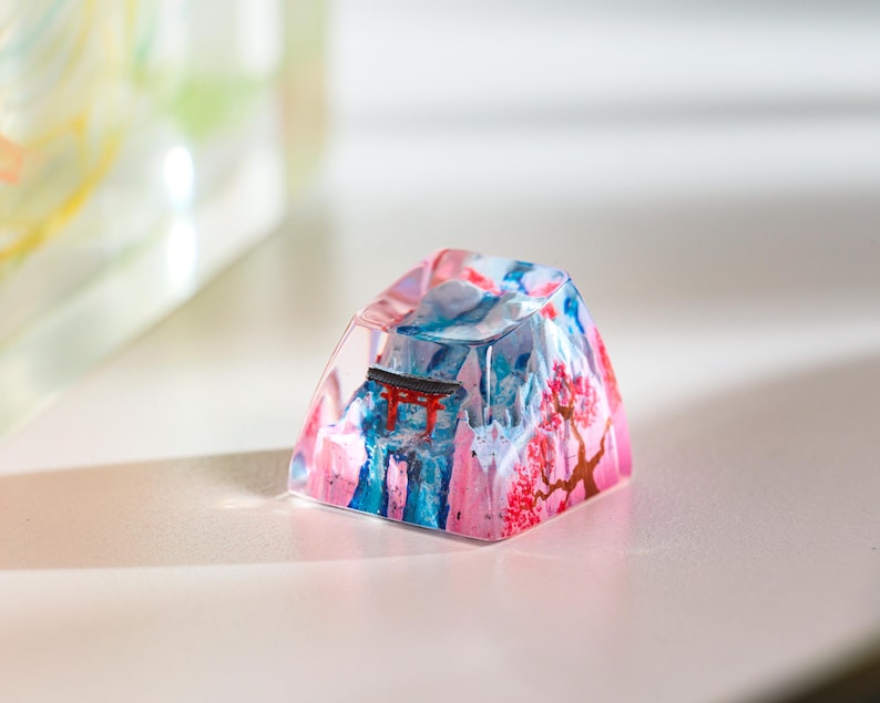 Puede incluir: Una tecla decorativa con un dise&ntilde;o de inspiraci&oacute;n japonesa. Presenta una escena de monta&ntilde;a con una puerta torii roja y flores de cerezo. La tecla est&aacute; hecha de resina y es una mezcla de rosa, azul y blanco. El dise&ntilde;o es art&iacute;stico.