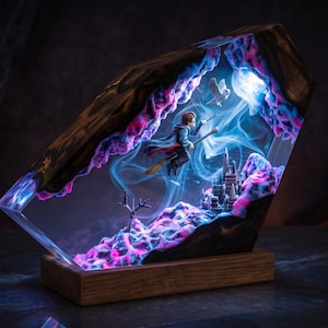 May include: A rectangular wooden lamp featuring a resin-encased diorama of a figure on a broomstick flying above a castle. The scene includes a white owl, blue smoke effects, and pink and blue textured elements, all illuminated from within.
