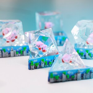 May include: Multiple clear resin sculptures in geometric shapes, each containing a pink character with red feet and a white face. The sculptures sit on a blue base with a cityscape design. The character is encased within the clear resin, giving a 3D appearance.