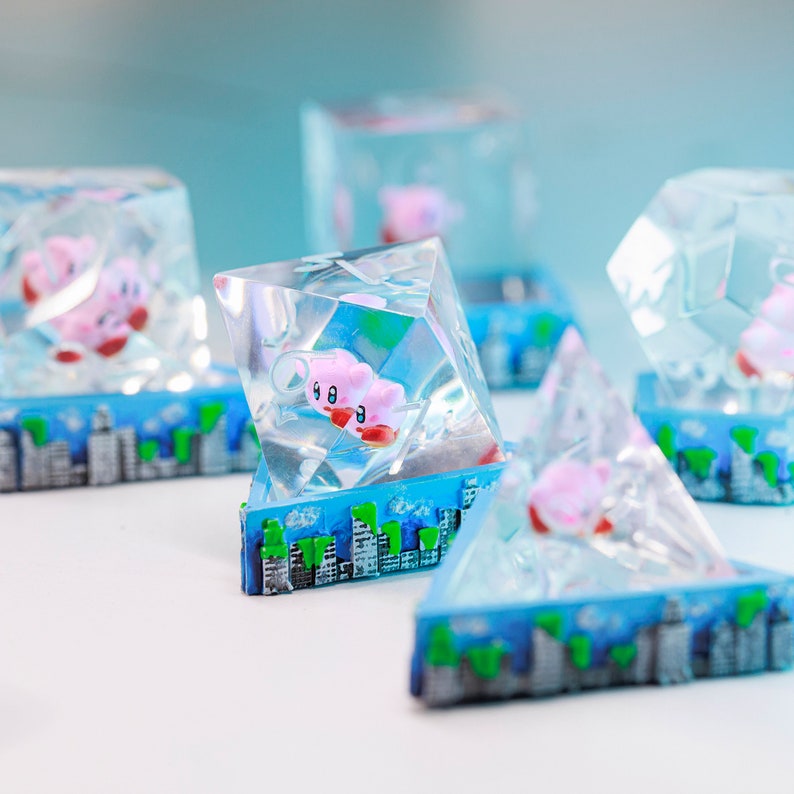 May include: Multiple clear resin sculptures in geometric shapes, each containing a pink character with red feet and a white face. The sculptures sit on a blue base with a cityscape design. The character is encased within the clear resin, giving a 3D appearance.