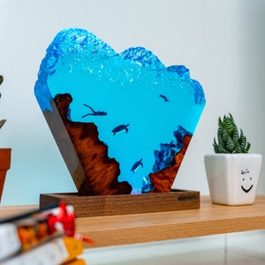 May include: A blue resin sculpture of an underwater scene with turtles and a diver. The sculpture is mounted on a wooden base and has a light inside. The sculpture is on a shelf with a potted plant and a book.