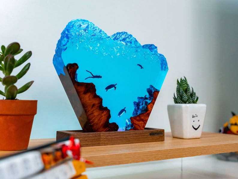 May include: A blue resin sculpture of an underwater scene with turtles and a diver. The sculpture is mounted on a wooden base and has a light inside. The sculpture is on a shelf with a potted plant and a book.