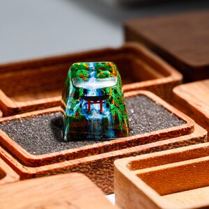 May include: A custom keycap featuring a scenic waterfall and Japanese torii gate design. The keycap is set in a wooden box with a dark gray foam insert. The keycap has a clear, glossy finish.