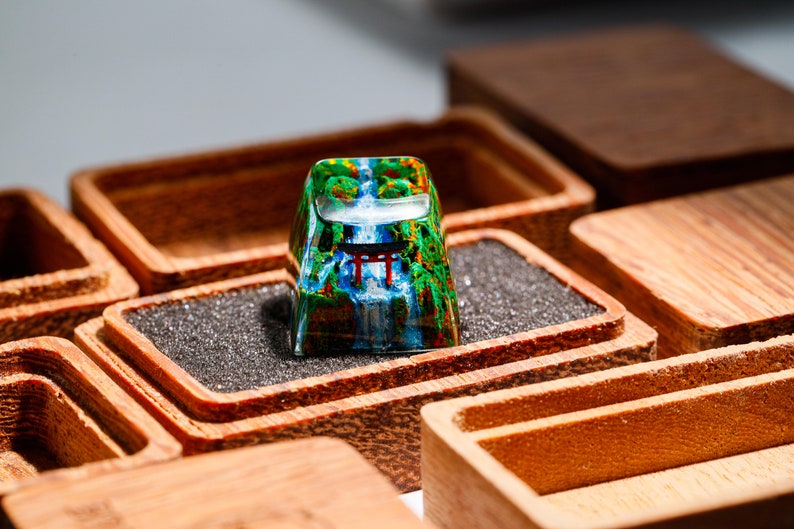 May include: A custom keycap featuring a scenic waterfall and Japanese torii gate design. The keycap is set in a wooden box with a dark gray foam insert. The keycap has a clear, glossy finish.