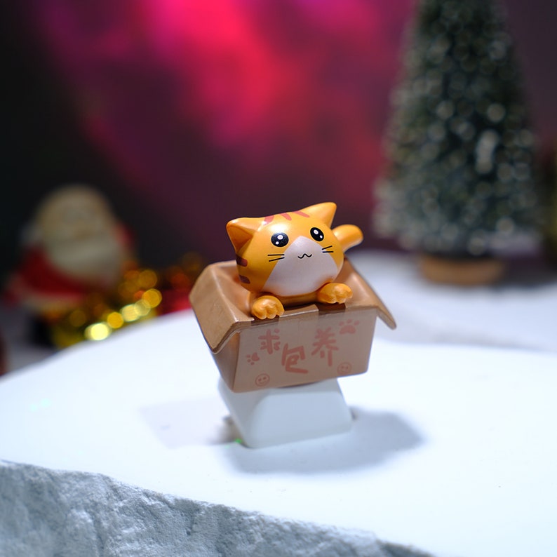 May include: A small, orange tabby cat figurine peeks out of a brown cardboard box. The box has Chinese characters printed on it. The cat has black eyes and is positioned on a white surface, with a blurred Christmas tree and Santa Claus in the background.