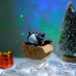 May include: A black and white cat figurine peeks out of a small cardboard box. The box has Chinese characters and paw prints. A small Christmas tree and a wrapped gift are in the background. The figurine is on a keyboard.