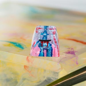 Puede incluir: Una tecla rectangular transparente con un dise&ntilde;o colorido. La tecla presenta una puerta torii roja, detalles en rosa, azul y blanco. La tecla es probablemente para un teclado mec&aacute;nico.