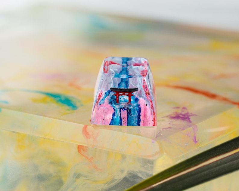Puede incluir: Una tecla rectangular transparente con un dise&ntilde;o colorido. La tecla presenta una puerta torii roja, detalles en rosa, azul y blanco. La tecla es probablemente para un teclado mec&aacute;nico.