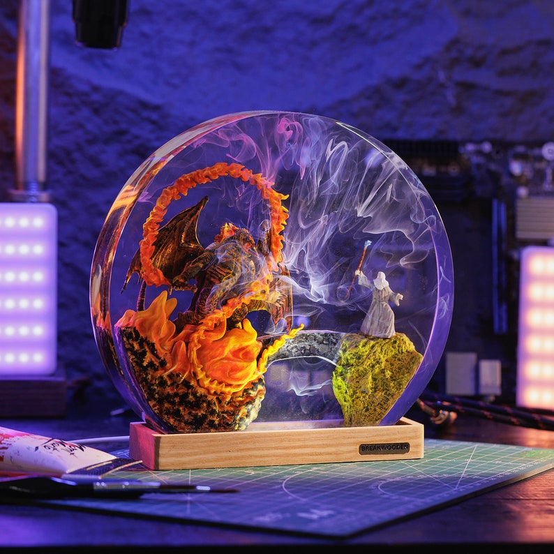 May include: A clear resin sphere with a dragon engulfed in orange flames on the left and a wizard on a rock on the right. The sphere sits on a wooden base, with a smoky effect inside. The scene is lit with purple and white lights.