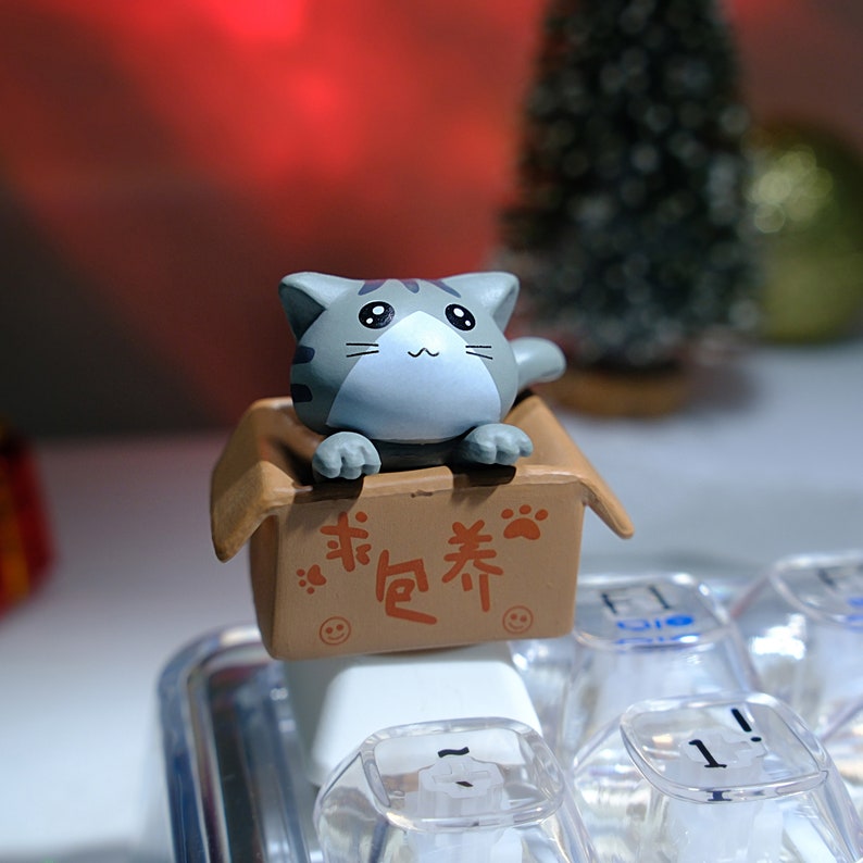 May include: A gray and white cat figurine peeks out of a brown cardboard box. The box has Chinese characters and paw prints. The figurine is placed on a keyboard with clear keycaps.