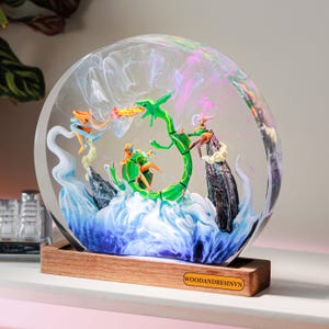 May include: A circular resin display featuring a green serpentine creature, a smaller orange figure, and a purple character amidst swirling white and blue clouds. The piece is mounted on a rectangular wooden base with the text WOODANDRESINVN.