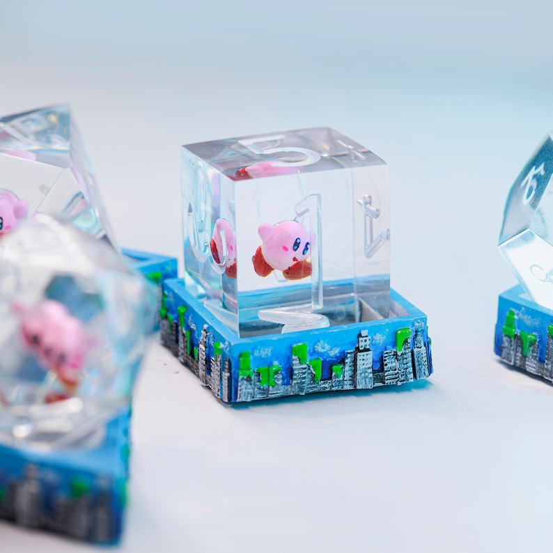 May include: Transparent resin cube featuring a pink Kirby figure, mounted on a blue base with a cityscape design. The cube has the number "1" etched on its side. Other similar cubes are visible in the background.