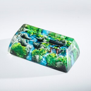 May include: A rectangular keycap with a detailed landscape design. The keycap features a miniature scene of a river flowing through a mountainous area with green foliage, blue water, and a small red and black structure.