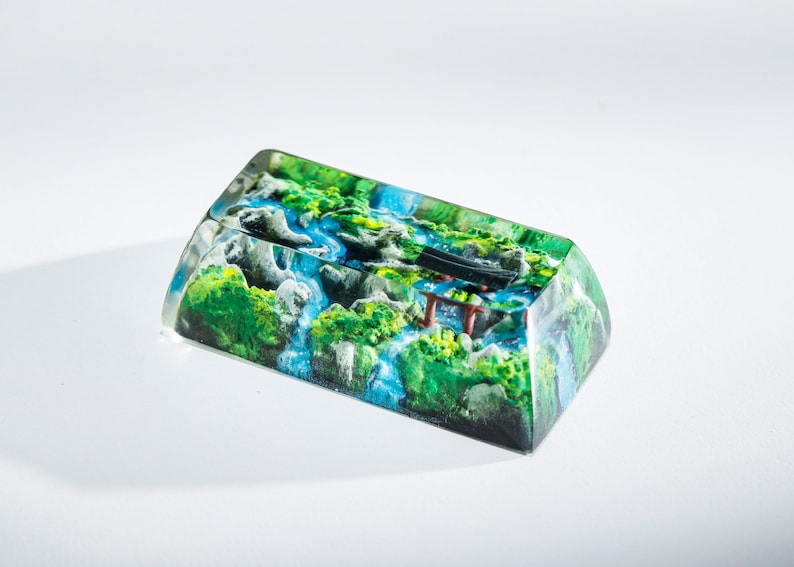 May include: A rectangular keycap with a detailed landscape design. The keycap features a miniature scene of a river flowing through a mountainous area with green foliage, blue water, and a small red and black structure.