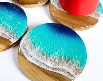 May include: Set of four round wooden coasters with a beach-themed design. Each coaster features a wooden base with a resin top depicting a beach scene with turquoise water and white foamy waves. A red mug rests on one coaster.