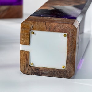 May include: A rectangular wooden block with a white panel and gold screws. The top of the block has a dark wood grain pattern with purple resin. Another similar block is in the background.