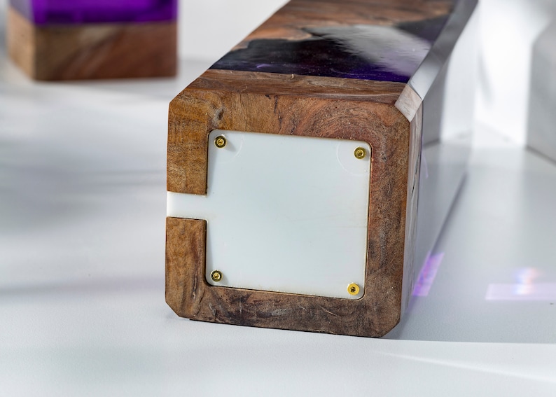 May include: A rectangular wooden block with a white panel and gold screws. The top of the block has a dark wood grain pattern with purple resin. Another similar block is in the background.