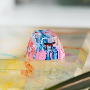 Puede incluir: Una colorida y art&iacute;stica tecla de resina con un dise&ntilde;o de inspiraci&oacute;n japonesa. La tecla muestra una puerta torii roja, flores de cerezo rosas y un paisaje monta&ntilde;oso azul y blanco. La tecla est&aacute; sobre una superficie multicolor.
