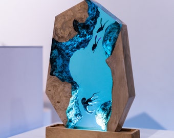 May include: A decorative wooden sculpture featuring a blue resin center with a diving scene. The sculpture has a natural wood frame and base. The resin depicts divers and an octopus. The sculpture is illuminated with blue light.