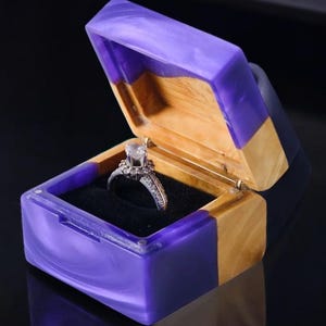 May include: A square ring box featuring a combination of natural light-toned wood and marbled purple resin. The box is open, revealing a black velvet interior lining that holds a silver-toned ring with a central clear stone and small side accents.