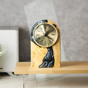 May include: A wooden desk clock with a gold-colored clock face and Roman numerals. The clock is set in a wooden base with a black resin design. The clock is displayed on a wooden shelf.