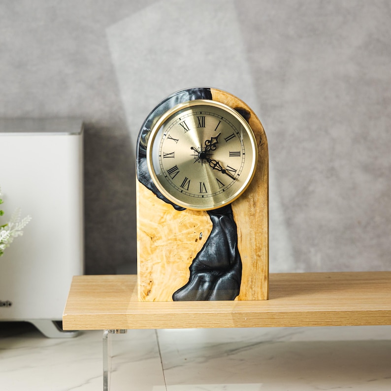 May include: A wooden desk clock with a gold-colored clock face and Roman numerals. The clock is set in a wooden base with a black resin design. The clock is displayed on a wooden shelf.