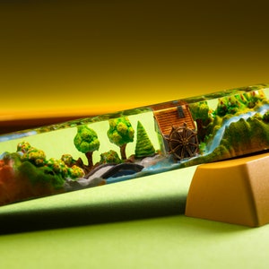 May include: A custom keyboard keycap featuring a miniature landscape scene. The keycap is clear with a green tint, showcasing a detailed diorama of a watermill, trees, and a river. The keycap is paired with a gold-colored key.