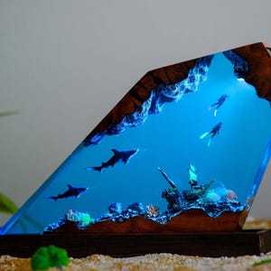 May include: A unique resin art piece depicting an underwater scene with sharks, divers, and a sunken ship. The artwork features a vibrant blue ocean, brown wooden accents, and a dark wooden base. The piece is illuminated with blue light.