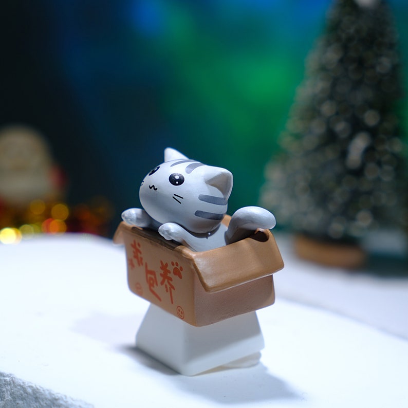 May include: A gray and white striped cat figurine peeks out of a brown cardboard box. The box has Chinese characters printed on it. The figurine is mounted on a white base, with a blurred green and blue background.