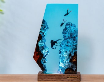 May include: A decorative resin lamp featuring a marine scene with divers and an octopus. The lamp has a wooden base and a blue and clear resin design. The lamp is a unique home decor item.