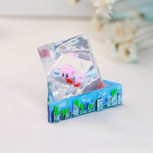 May include: A clear resin sculpture in a geometric shape, with a pink cartoon character inside. The base is a blue box with a cityscape design, featuring white buildings and green trees. The sculpture is a decorative item.
