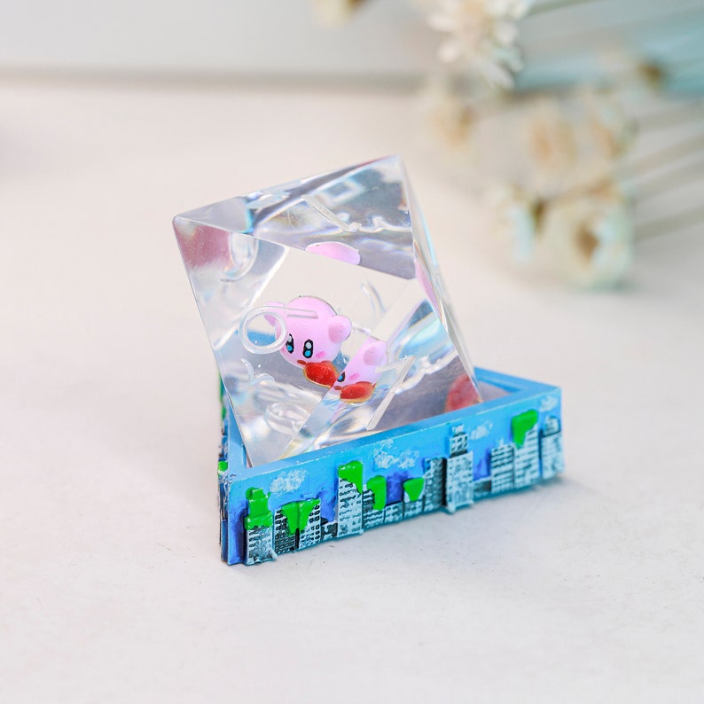May include: A clear resin sculpture in a geometric shape, with a pink cartoon character inside. The base is a blue box with a cityscape design, featuring white buildings and green trees. The sculpture is a decorative item.