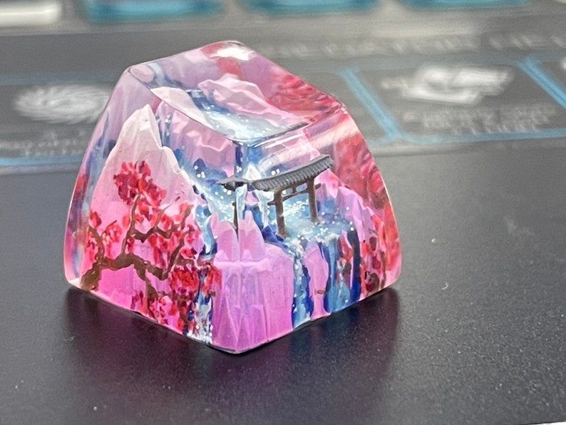 May include: A clear pink and blue resin keycap with a miniature Japanese-style temple and a cherry blossom tree painted inside. The keycap is designed to look like a mountain scene with a waterfall.