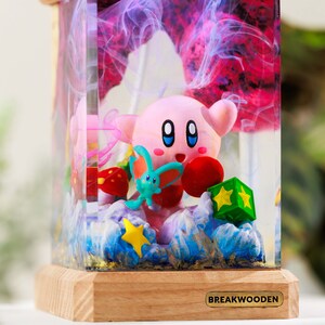 May include: A rectangular resin lamp featuring a pink Kirby figure, a small teal creature, a green star block, and a yellow star, all encased in clear resin with swirling purple and pink smoke effects, set on a light wood base with a metal tag reading BREAKWOODEN.