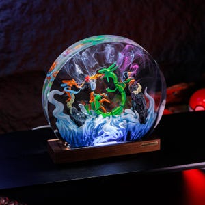 May include: A circular resin lamp featuring a green serpentine creature and orange figures amidst swirling white and blue smoke effects, set on a rectangular wooden base with the text BREAKWOODEN engraved on a small metal plate on the front.
