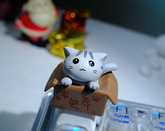 May include: A small, gray and white cat figurine sits inside a brown cardboard box with Chinese characters. The box is on a white keyboard with blue lights.