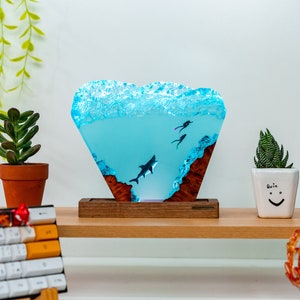 May include: A blue resin sculpture of a shark and two divers, set on a wooden base. The sculpture is shaped like a wave and has a light blue color. The base is made of dark wood and has a natural wood grain.