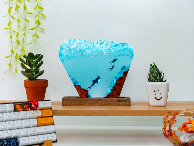 May include: A blue resin sculpture of a shark and two divers, set on a wooden base. The sculpture is shaped like a wave and has a light blue color. The base is made of dark wood and has a natural wood grain.