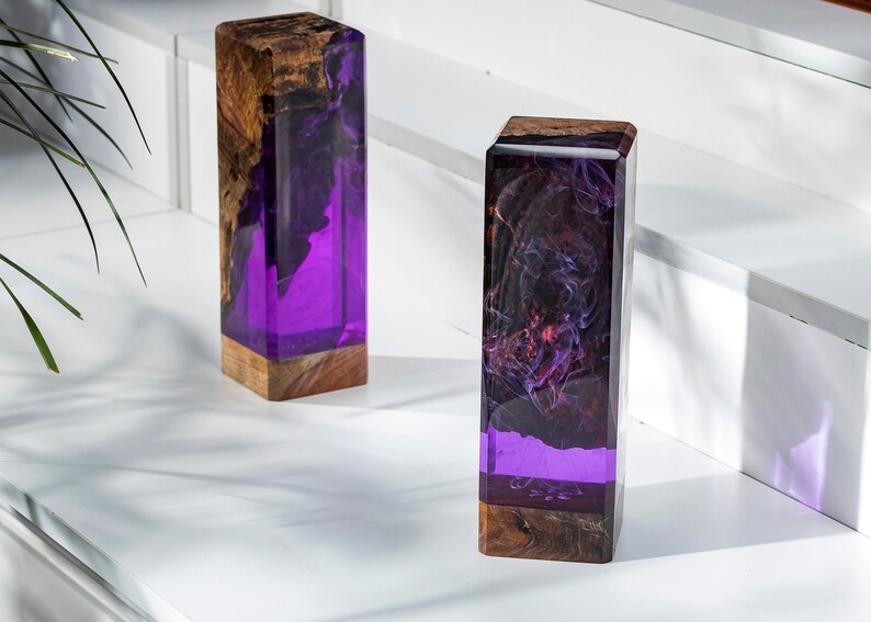 May include: Two rectangular decorative blocks. The base is a natural wood grain, transitioning to a vibrant purple resin. The resin is swirled with dark, smoky patterns. The blocks are displayed on a white surface.