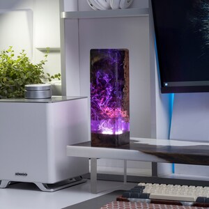 May include: A rectangular wooden and resin lamp with a vibrant purple and pink swirling design. The lamp is on a white desk with a computer monitor and keyboard in the background. The lamp is approximately 10 inches tall.
