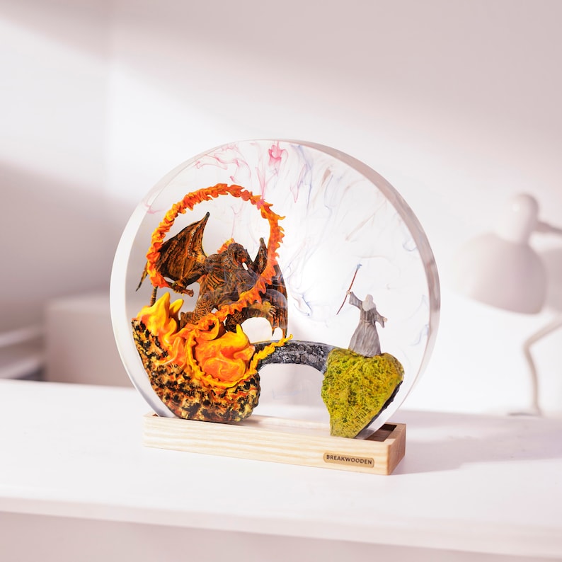 May include: A resin sculpture featuring a dragon engulfed in flames facing a wizard with a staff. The scene is encased in a clear, circular resin with colorful swirls, set on a wooden base. The base has the text "BREAKWOODEN".