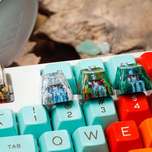 Puede incluir: Primer plano de un teclado mec&aacute;nico con teclas personalizadas. Las teclas presentan dise&ntilde;os de paisajes coloridos con cascadas y puertas Torii japonesas. El teclado tiene una base blanca con teclas de colores.