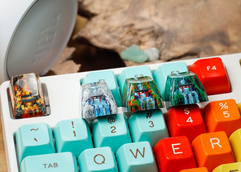 Puede incluir: Primer plano de un teclado mec&aacute;nico con teclas personalizadas. Las teclas presentan dise&ntilde;os de paisajes coloridos con cascadas y puertas Torii japonesas. El teclado tiene una base blanca con teclas de colores.