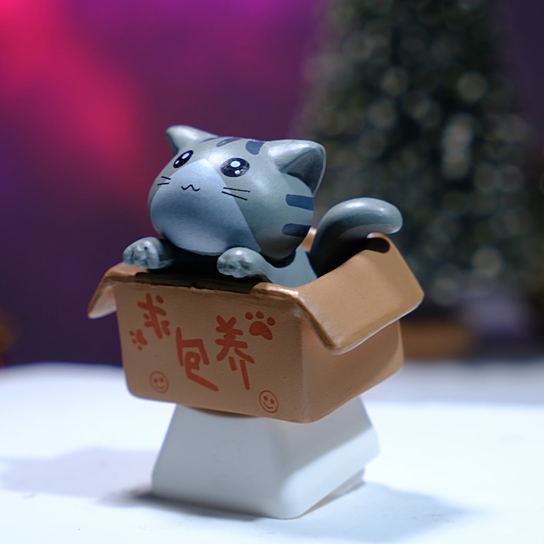 May include: A gray tabby cat figurine peeks out of a small brown cardboard box. The box has red Chinese characters and paw prints. The cat and box are mounted on a white rectangular base. The background is blurred with pink and green hues.