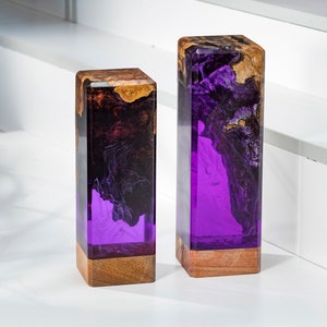 May include: Two rectangular decorative blocks. The top portion is a mix of brown and gold wood, while the lower portion is a vibrant purple resin. The blocks are set on a white surface.